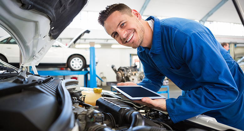 A male student in a garage is dressed in blue overalls. He holds a tablet and observes a car with a positive expression.
