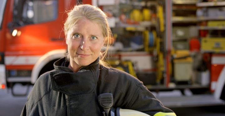 A female firefighter poses confidently in her uniform looking directly into the camera. She stands beside a fire truck, ready for action.