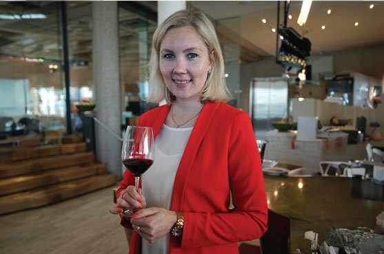 Kati Vainionpaa is in a bar space looking at the camera. She wears a red blazer over a cream top and holds a glass of red wine.
