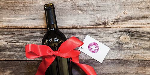A wine bottle against a background of unpainted wooden boards, tied with a red ribbon and with a gift card and the stencil of a lipstick kiss.