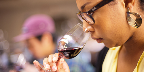 A woman holds a glass to smell red wine, as part of tasting, called "nosing" the wine. She wears a pale yellow top, has her hair tied back to reveal gold earrings and wears dark rimmed glasses.