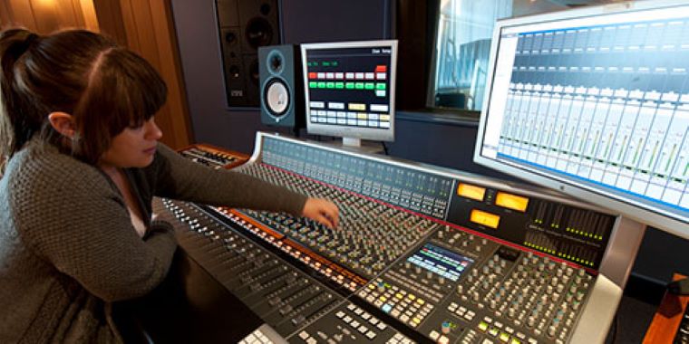 A woman is working in a professional recording studio at an audio mixing console.  She has multiple screens in front of her displaying audio waveforms as she adjusts sound levels on a large soundboard. 
