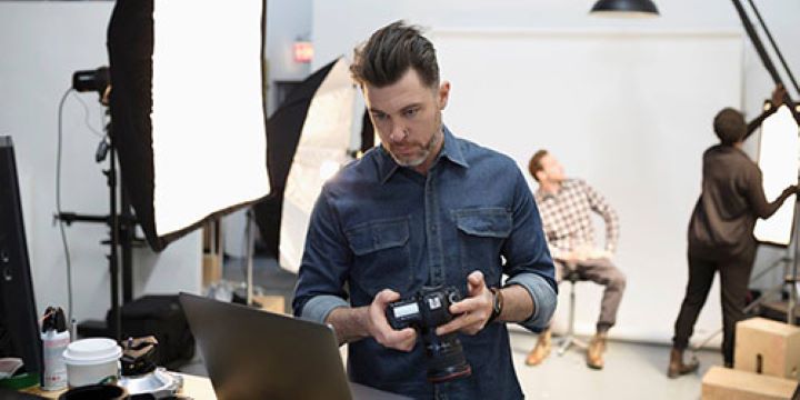 A man in a denim shirt and slicked back brown hair is holding a digital camera while looking at a laptop screen in a photography studio. There are people working behind him. One is seated in front of a white screen while the other adjusts overhead studio lighting. 