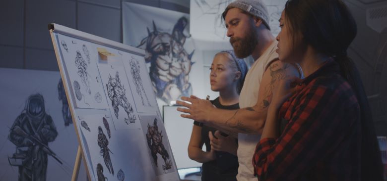 A group of three artists collaborate on an art project, discussing detailed sketches of characters on a whiteboard. The sketches feature fantasy and sci-fi-themed designs, including armoured figures and futuristic soldiers.