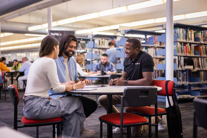 TAFE NSW staff member providing library learning support to two students at a circular table, with bookshelves and other learners in the background.
