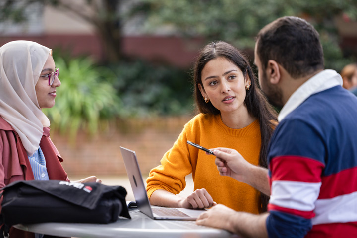 Three students sitting outdoors around a table, discussing and collaborating with a laptop and notes.