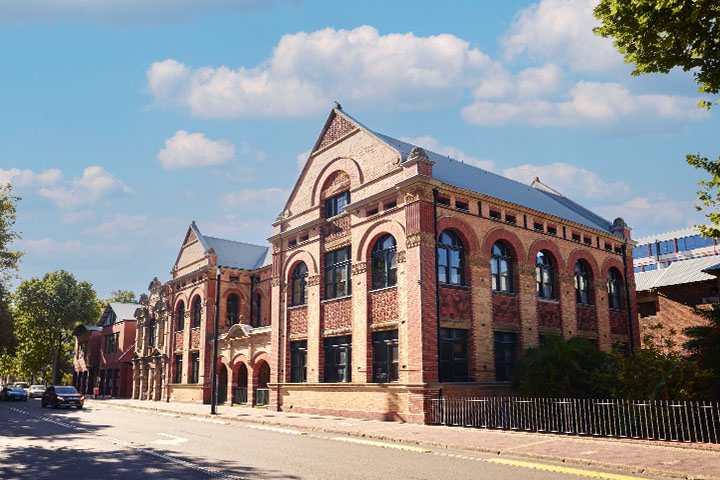 TAFE NSW Hunter Street campus in Newcastle featuring a heritage-style brick building with ornate detailing, large arched windows, and pitched roofs, situated along a quiet street lined with trees under a partly cloudy blue sky.