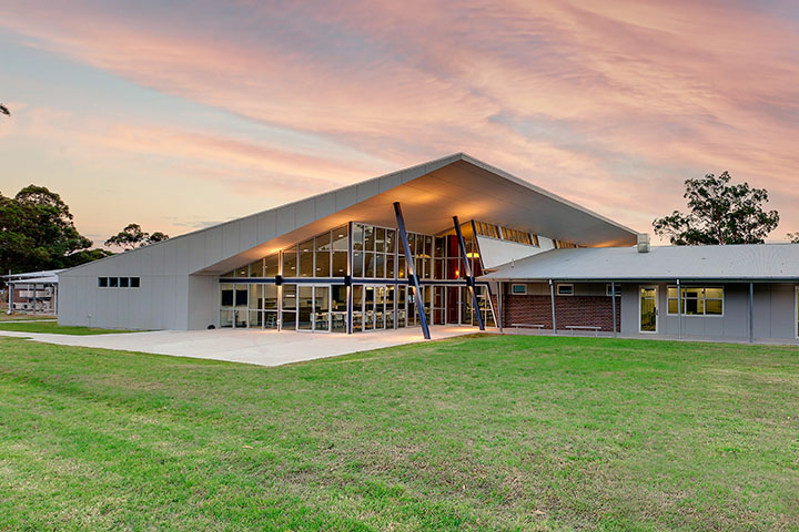 Contemporary TAFE NSW Kurri Kurri campus building with large glass windows, angled roofline, and steel supports, set against a vibrant pink and orange sunset sky with green lawn in the foreground.