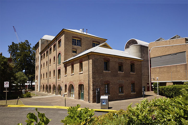 TAFE NSW Hamilton campus featuring multiple heritage‑style brick buildings with arched windows and metal roofing, arranged around a paved courtyard with greenery in the foreground, set against a clear blue sky.