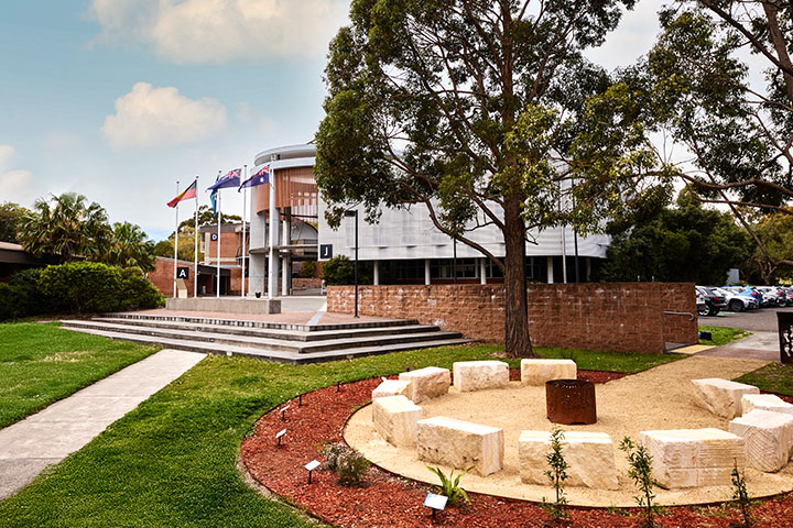 TAFE NSW Shellharbour Campus featuring a modern building with large windows, multiple flags at the entrance, and a landscaped outdoor area with circular stone benches surrounded by greenery.