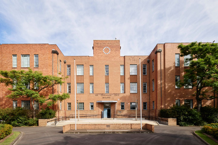 Historic TAFE NSW Tighes Hill campus building with symmetrical red-brick façade, central entrance ramp, tall windows, and two flagpoles in front, surrounded by green trees under a bright blue sky with scattered clouds.