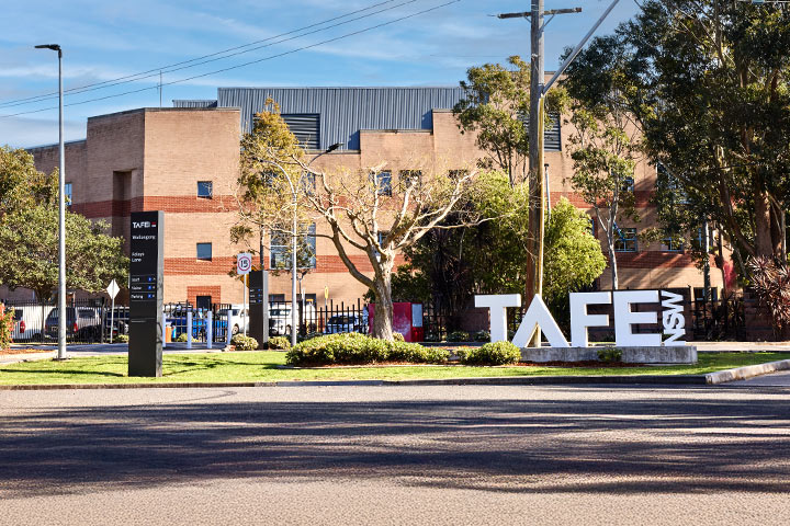 TAFE NSW Wollongong campus featuring a large brick building with multiple windows, surrounded by trees and greenery, with prominent campus signage in the foreground under a partly cloudy sky.