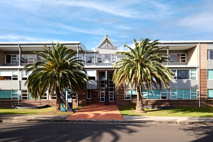 TAFE NSW Wollongong West campus building with large windows and a central entrance, flanked by two tall palm trees and a red pathway, set against a partly cloudy sky and green lawn.
