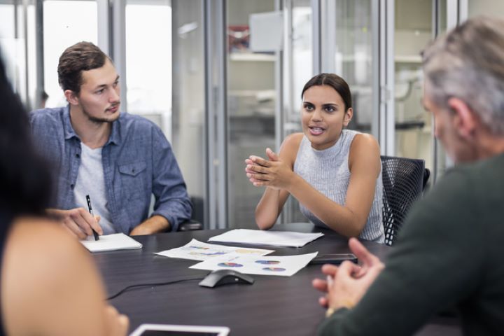 An Aboriginal woman sits at a corporate desk with colleagues listening to her intently. There are pieces of paper scattered on the dark wood desk and glass walls and doors of other study spaces, in the background.