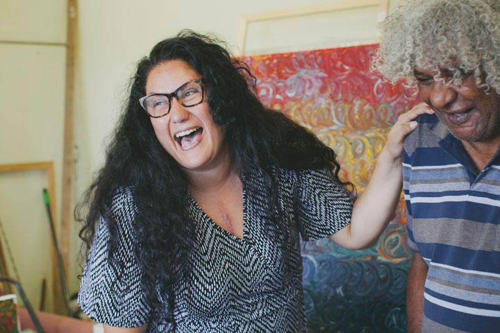 Two people sharing a joyful moment: a woman with long curly hair and glasses laughs heartily, while a man with curly gray hair smiles warmly, standing together in a vibrant, art-filled room.