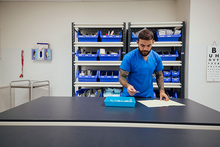 An Aboriginal medical worker wearing blue scrubs stands in a laboratory at a table. He is placing a sticker on a form, and there is pathology equipment on shelves behind him.