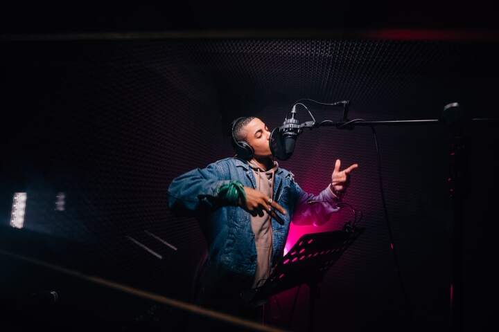 A student sings with closed eyes into a microphone in a dimly lit recording studio. The person wears headphones and makes expressive hand movements, showing that they’re in the flow.