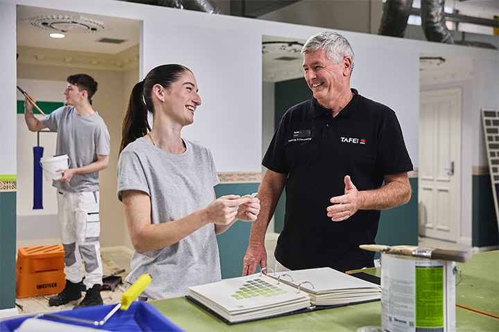 Painting student and teacher discuss colour selections in a training workshop, with paint samples open on the table and another student applying paint to a wall in the background.