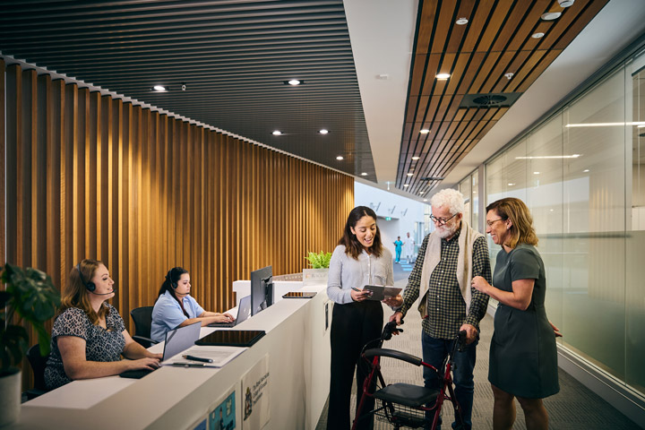 Reception area with two staff working on computers and three people interacting, including one with a tablet assisting an elderly person using a walker.