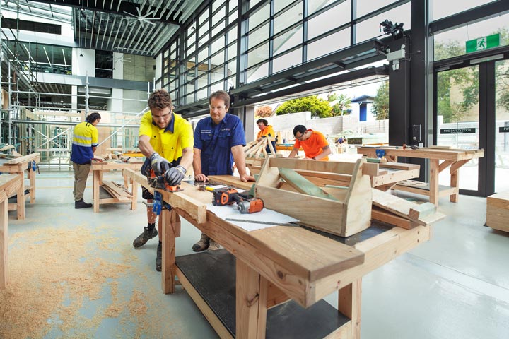 A male student, guided by his teacher, works on a carpentry project in a workshop. Two other students work on their projects in the background.