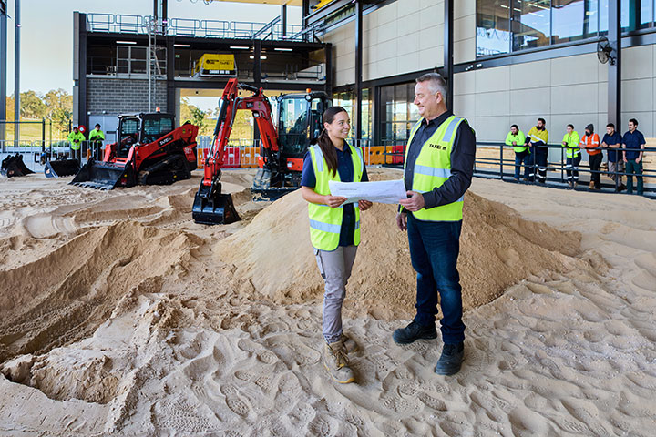 A female student works with the teacher in a civil construction workshop with a bulldozer and excavator in the background.
