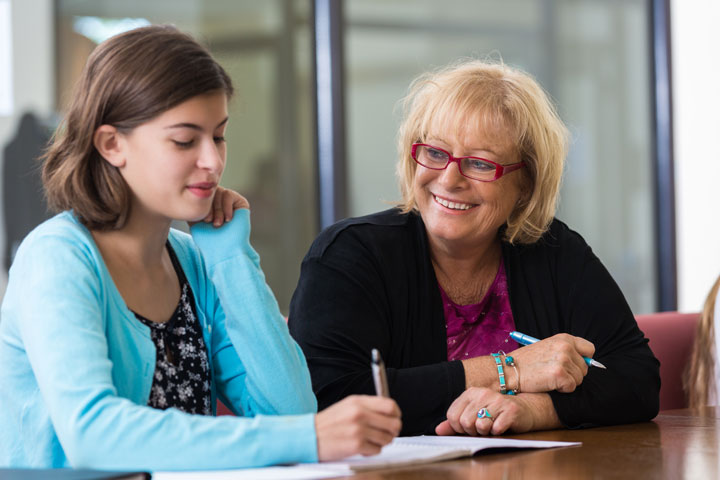A teacher with short blond hair and glasses is sitting smiling beside the student next to her. The student is holding a pen and writing in her workbook.