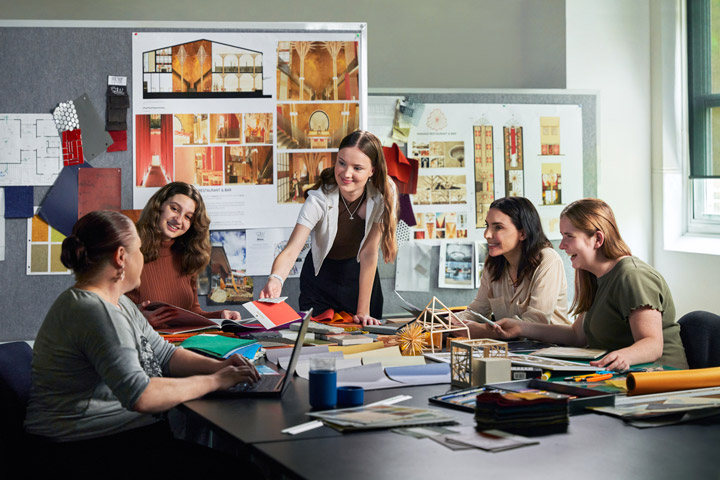 Five women working together in a design studio engaged in discussion. They are gathered around a large table covered with design materials such as fabric samples, architectural models, and colour swatches. In the background, there are various design plans, sketches and inspiration boards pinned to a grey wall.