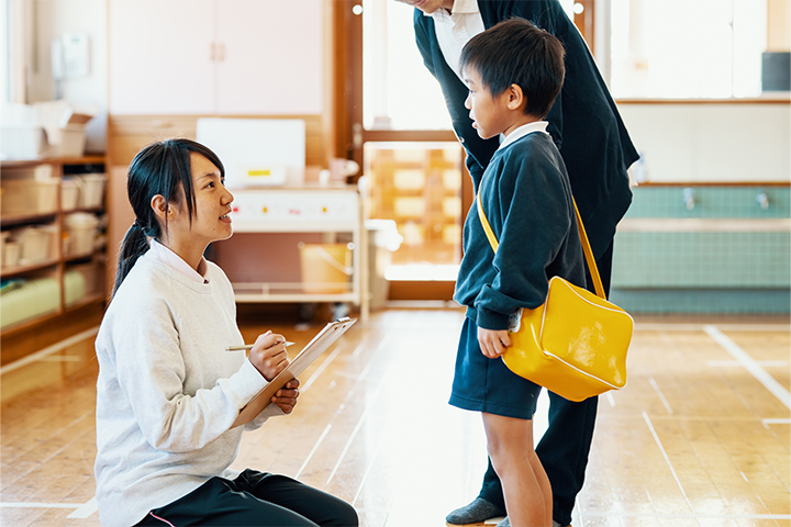 A teacher talks to a small boy in a school uniform, holding a yellow bag around his shoulder