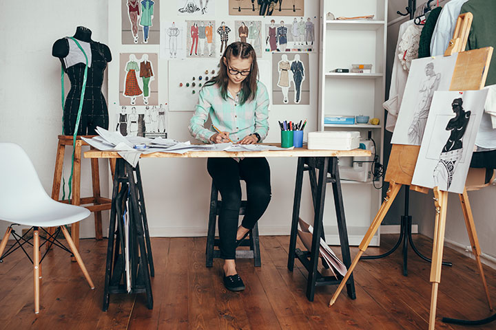 A young woman sits at a desk covered in papers and drawings. She’s working on some sketches. There are art easels with graphics around her, fashion sketches pinned on the wall and a mannequin beside her.