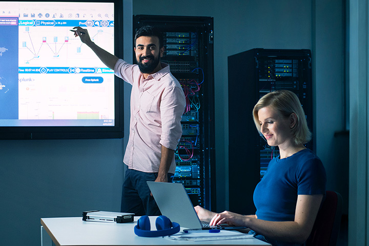 A cyber security teacher pointing at a digital board while a student works on a laptop in a classroom setting.