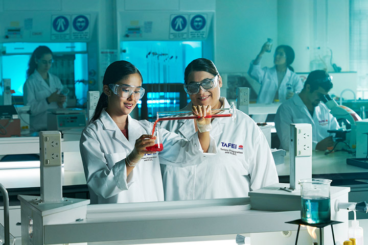 Two female students wearing safety glasses and lab coats collaborate on a science experiment in a bright laboratory classroom. They carefully measure and mix colorful chemicals in beakers, while other students work diligently at nearby tables.