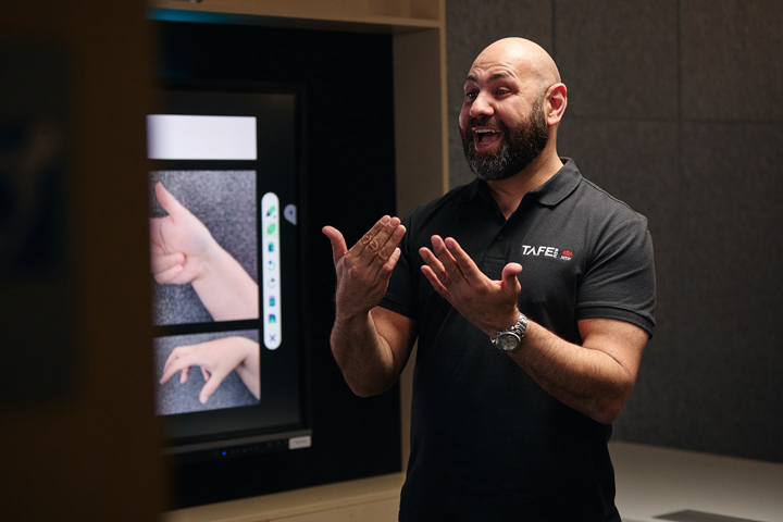 A man with a beard wearing a TAFE NSW polo shirt is using sign language while standing in front of a screen displaying images of hands making different signs. The man appears to be engaged and expressive as he communicates.