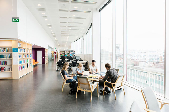 A group of students are studying together at a table in a bright, modern library with large windows overlooking a cityscape. Shelves of books and computer workstations line the open space.