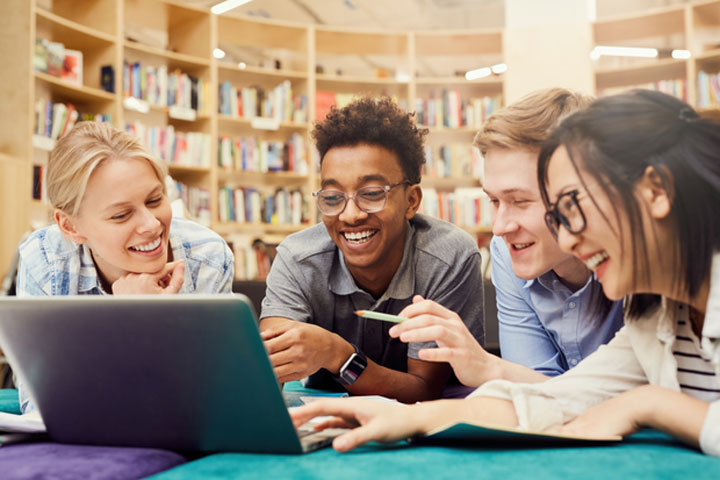 A diverse group of four young adults, consisting of two men and two women, are gathered around a laptop in a library. They are all smiling and appear to be enjoying their collaborative work or study session. The background features bookshelves filled with books.