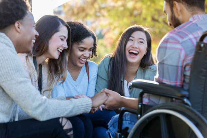 A diverse group of young adults, including one in a wheelchair, laughing closely together while holding hands outdoors.
