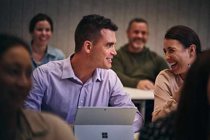 A student with his laptop open engaging with a smiling fellow student in a face-to-face class.
