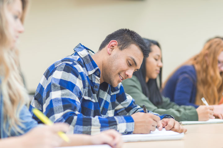 A man is working besides his classmates with a pen and paper in hand. He is writing notes and is wearing a blue and white checkered shirt. He is smiling and looks pleased.