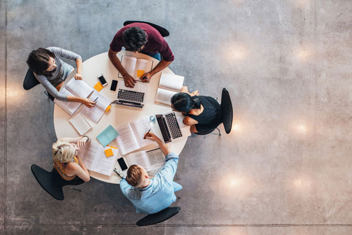 A birds-eye-view image of a group of students sat around a round table, studying. There are open books, laptops and notes on the table. The environment appears collaborative. 