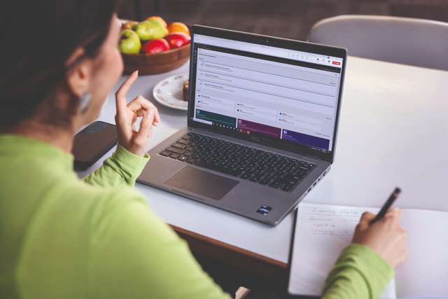 A woman sits at her desk with her computer open, studying online. She’s writing notes and appears to be smiling.