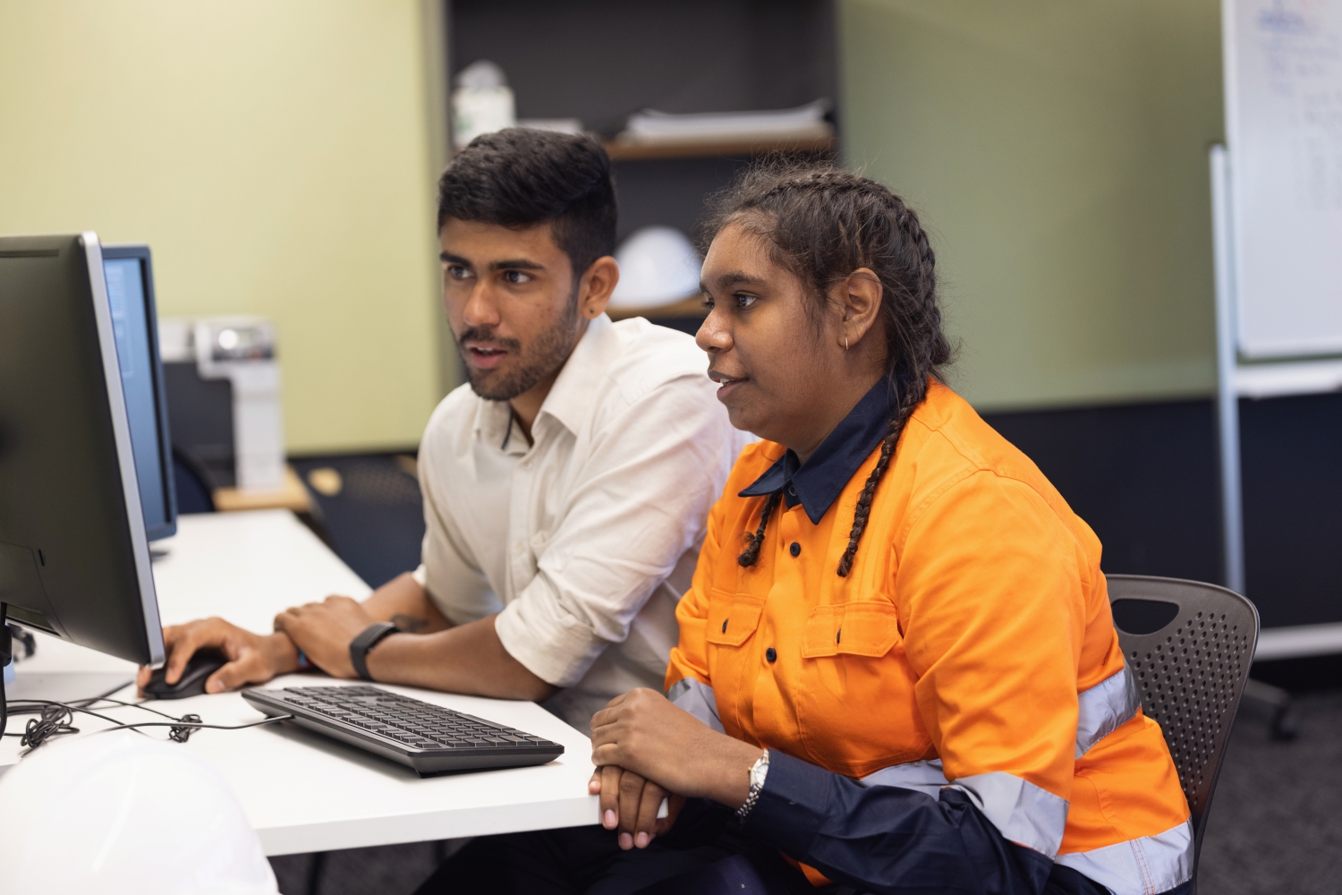 Two people are sitting at a computer. One person is showing the other person something on the screen.