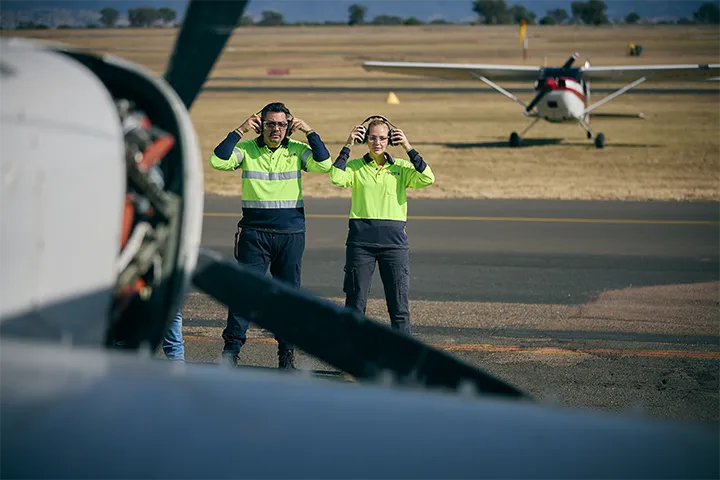 Two aviation students in high-visibility shirts stand on an airfield, adjusting their headsets as they prepare for aircraft operations. A small airplane is visible in the background.