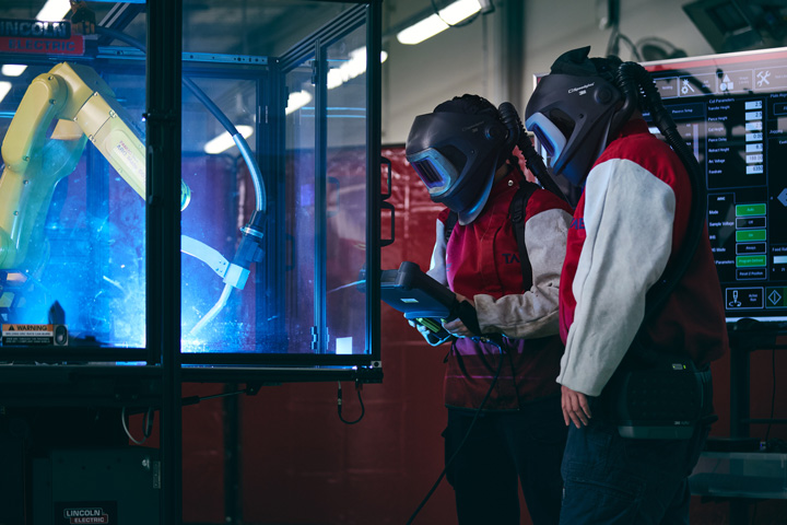 Two TAFE NSW students wearing welding helmets and protective gear monitor and control a robotic welding arm inside a safety enclosure, using a digital control panel as part of advanced robotic welding training at a Centres of Excellence facility.