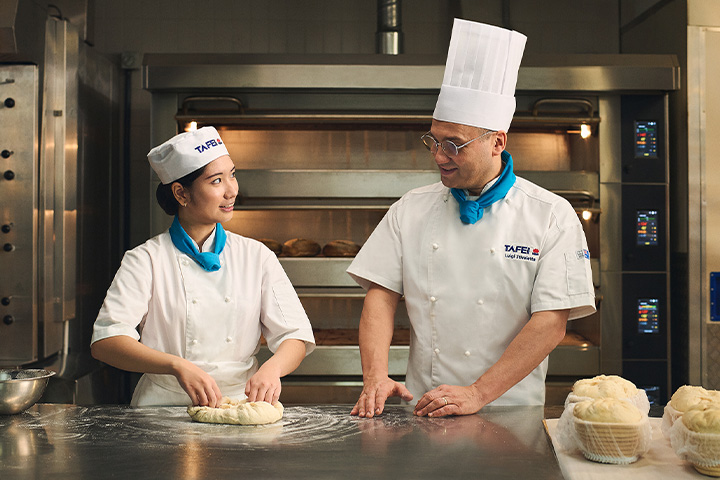 A baking student and their instructor share a smile while kneading dough during a bread-making lesson.