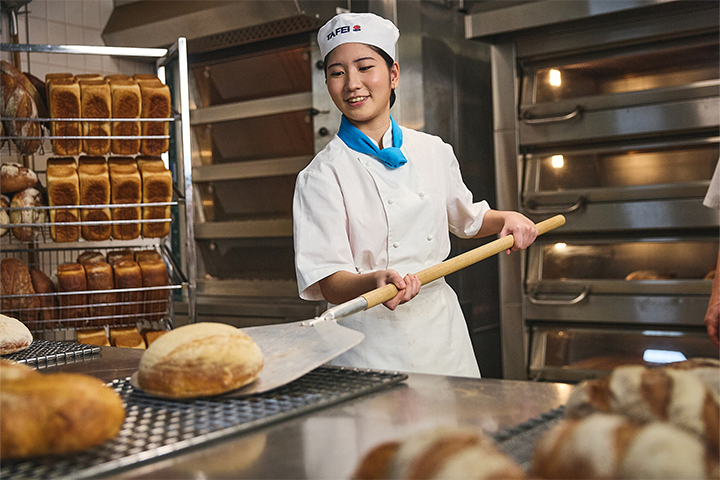 A female baker in a white TAFE NSW chef's uniform is shaping handling freshly-baked bread straight from a commercial kitchen oven.