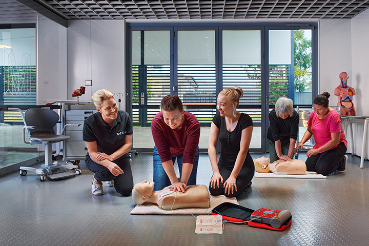 A teacher leads a first aid class, demonstrating CPR on a dummy with adult students.