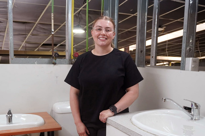 Angel Bielefeld stands in a partially constructed bathroom. She is wearing a black t-shirt, clear glasses, and smiles at the camera.