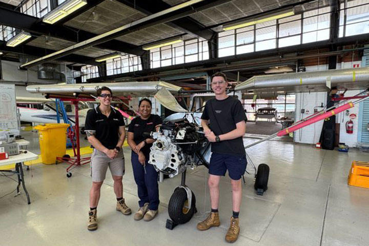 Three people stand in front of a small aircraft under construction in an aviation workshop. They are smiling and wearing safety glasses. The workshop contains tools, equipment, and parts of the aircraft, including its propeller and partially exposed engine.