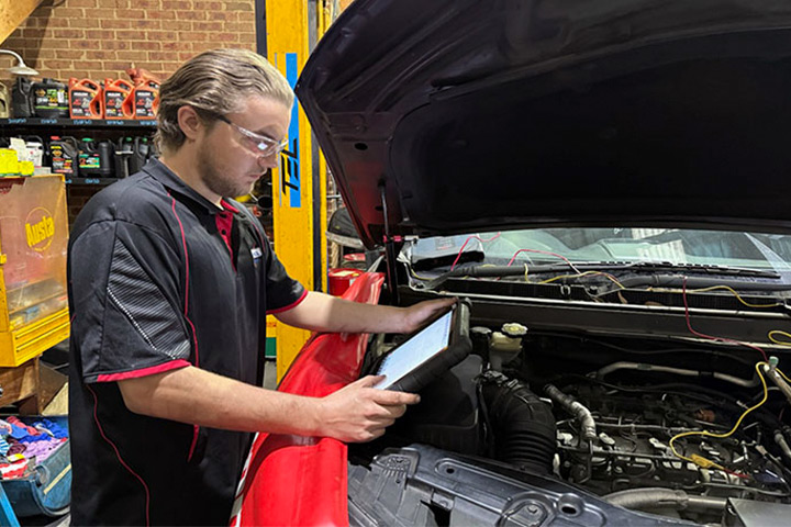 A mechanic holding a diagnostic tablet while inspecting the engine of a car in a garage. The background includes tools, shelves with containers, an automotive lift, and mechanical parts.