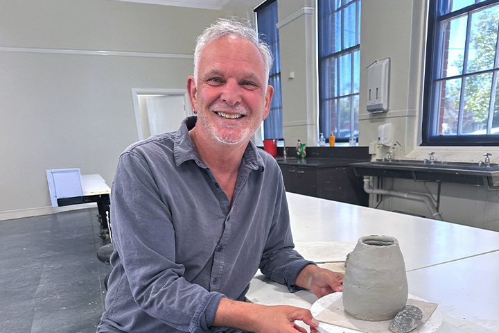 TAFE NSW Dubbo Visual Arts teacher Garry Jones sits at a workbench smiling with a clay sculpture in the shape of a vase.