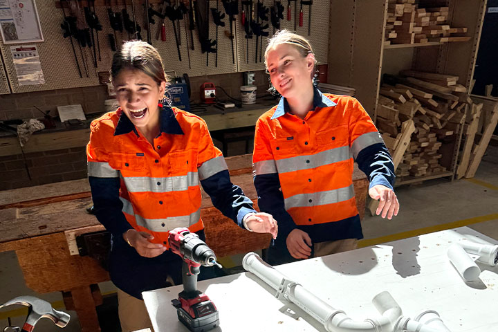 St Peters year 10 students Sienna Bantock and Poppy Melgaard pictured at a workbench, reaching for a drill and plumbing pipes. They wear orange high visibility work shirts and appear to be laughing.